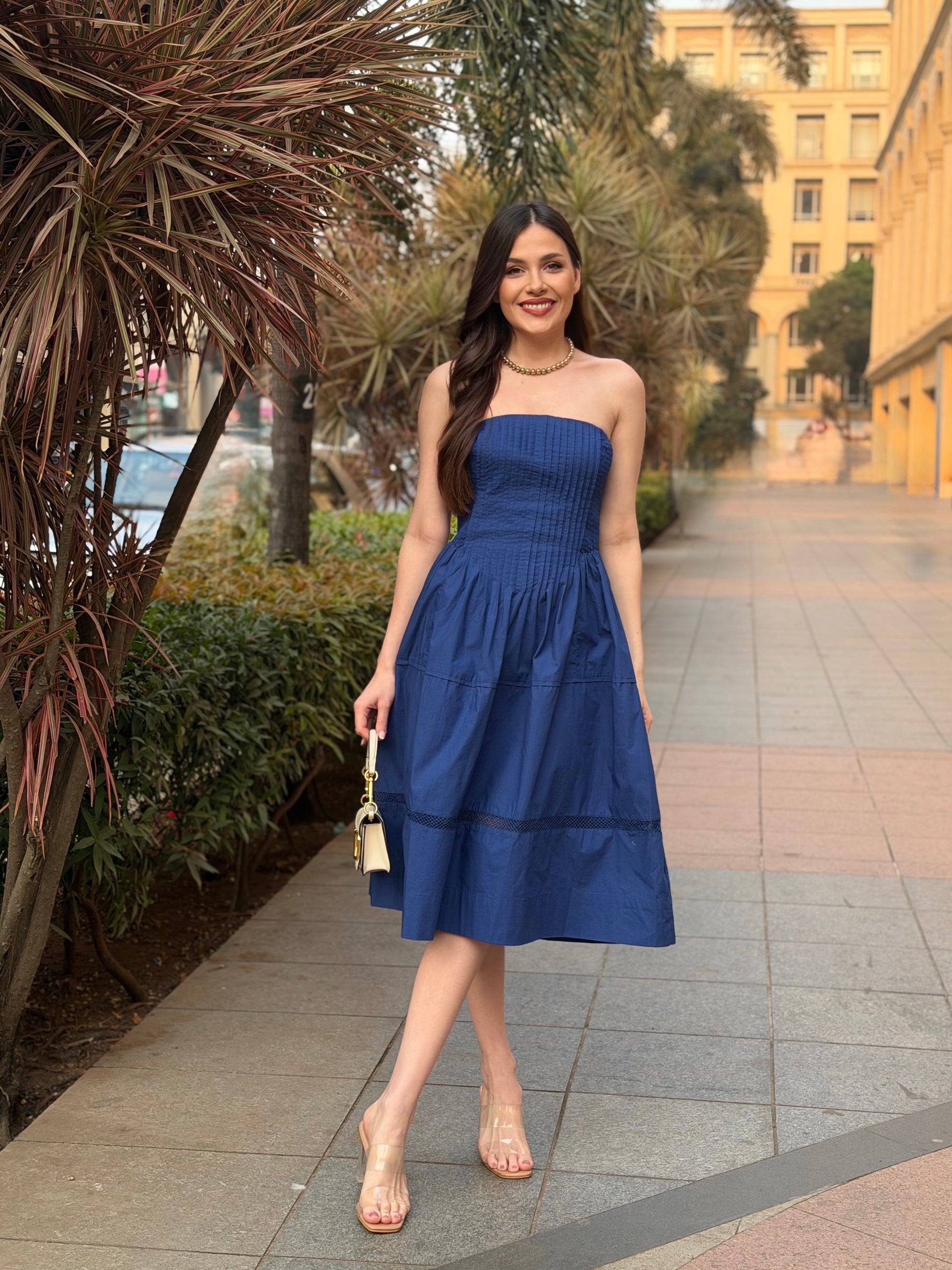 Woman in a blue dress standing on a sidewalk with greenery and a building in the background