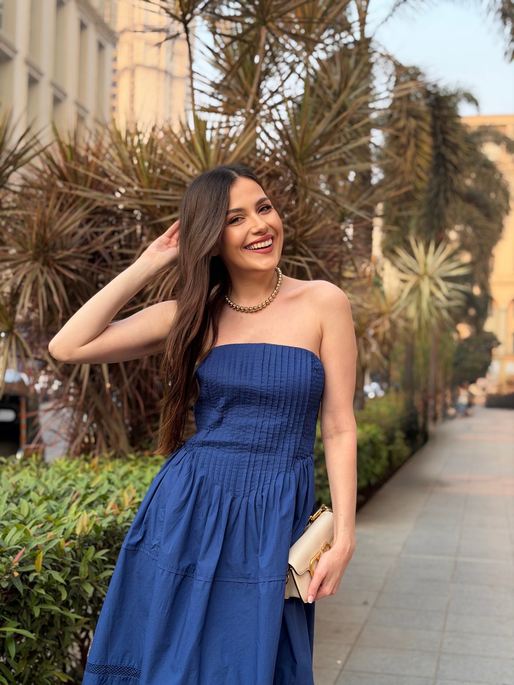 Woman in a blue strapless dress standing outdoors with plants and buildings in the background, western dress in india, 