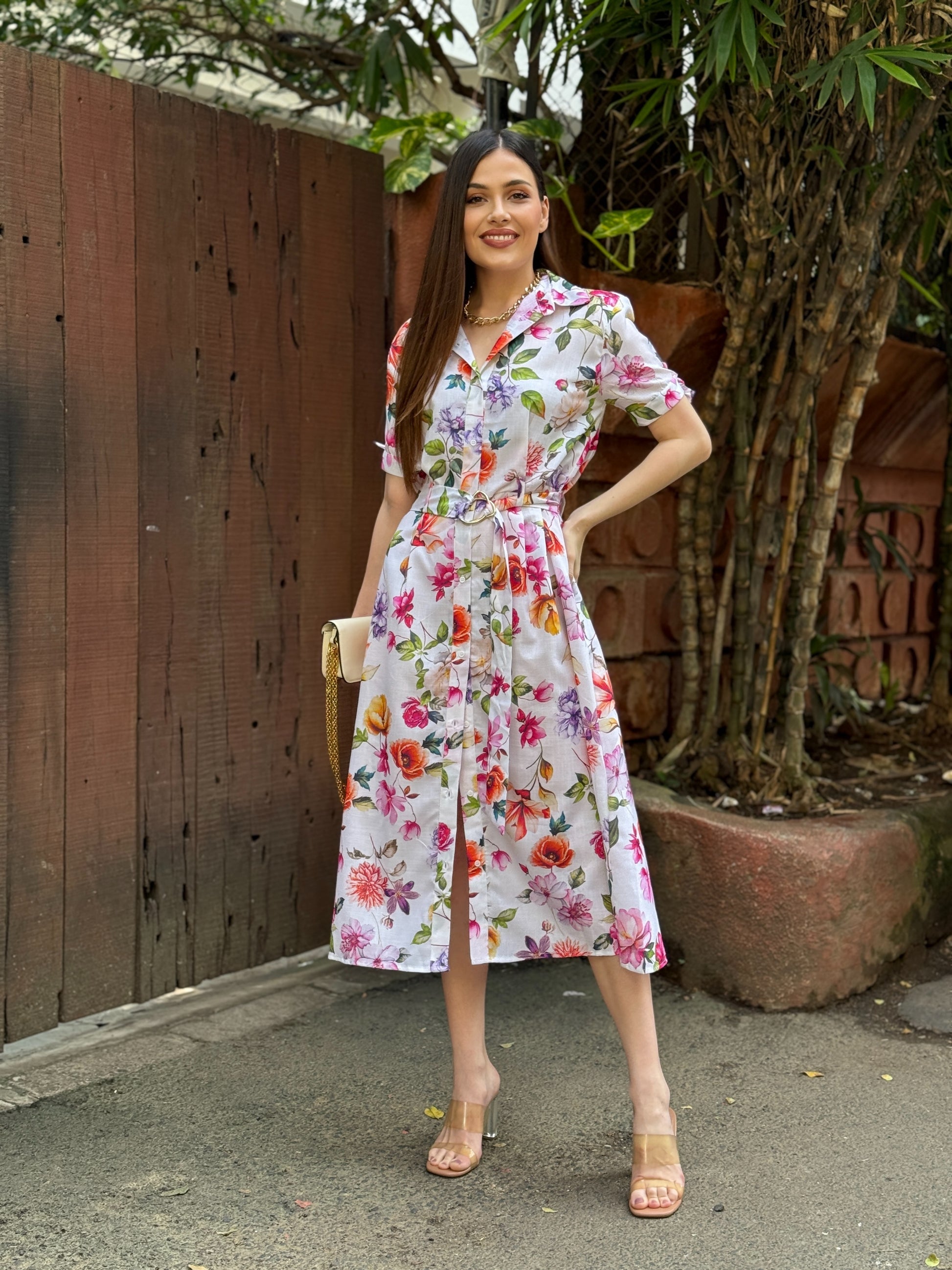 Woman wearing a floral dress standing outdoors next to a wooden fence and tree.